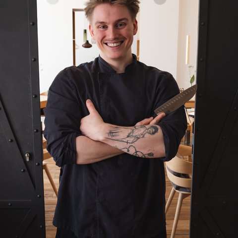 Restaurant in Parsberg: Enjoy gourmet Young chef in black outfit holding knife, smiling with tattoo on arm
